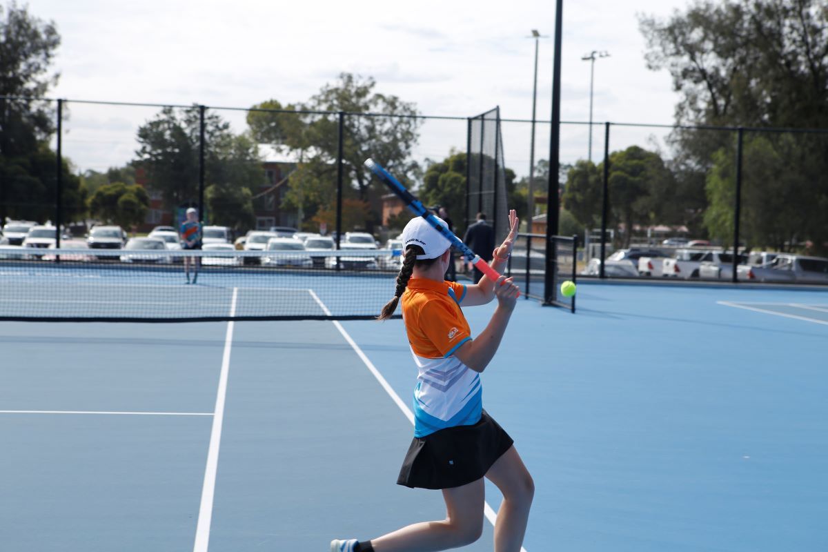 A young girl aged approximately ten is swinging a tennis racquet in preparation to hit a yellow tennis ball in flight. She is dressed in brightly coloured sporting gear and running across a new blue tennis court.