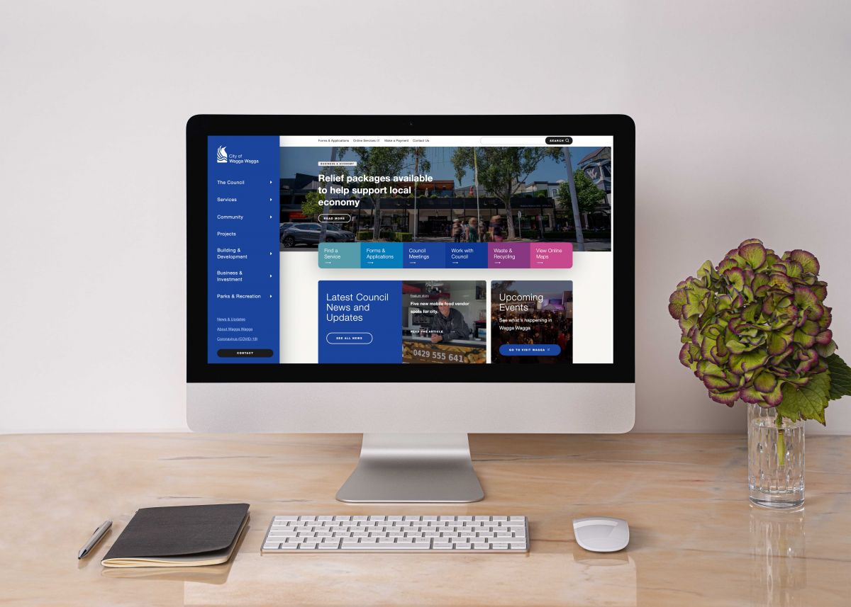 A computer monitor showing Wagga Wagga City Council's new homepage sits on a desk with a notepad, pen, keyboard, mouse and vase of flowers.
