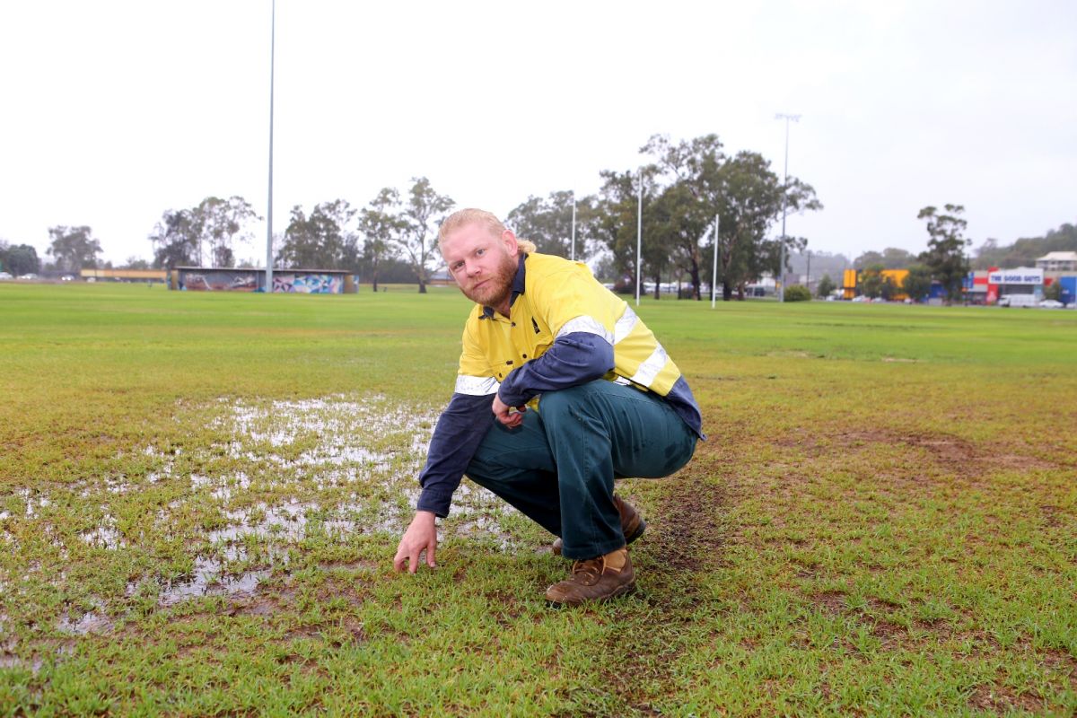 Man in high visibility jumper crouches down to test the wet ground at a sporting field 