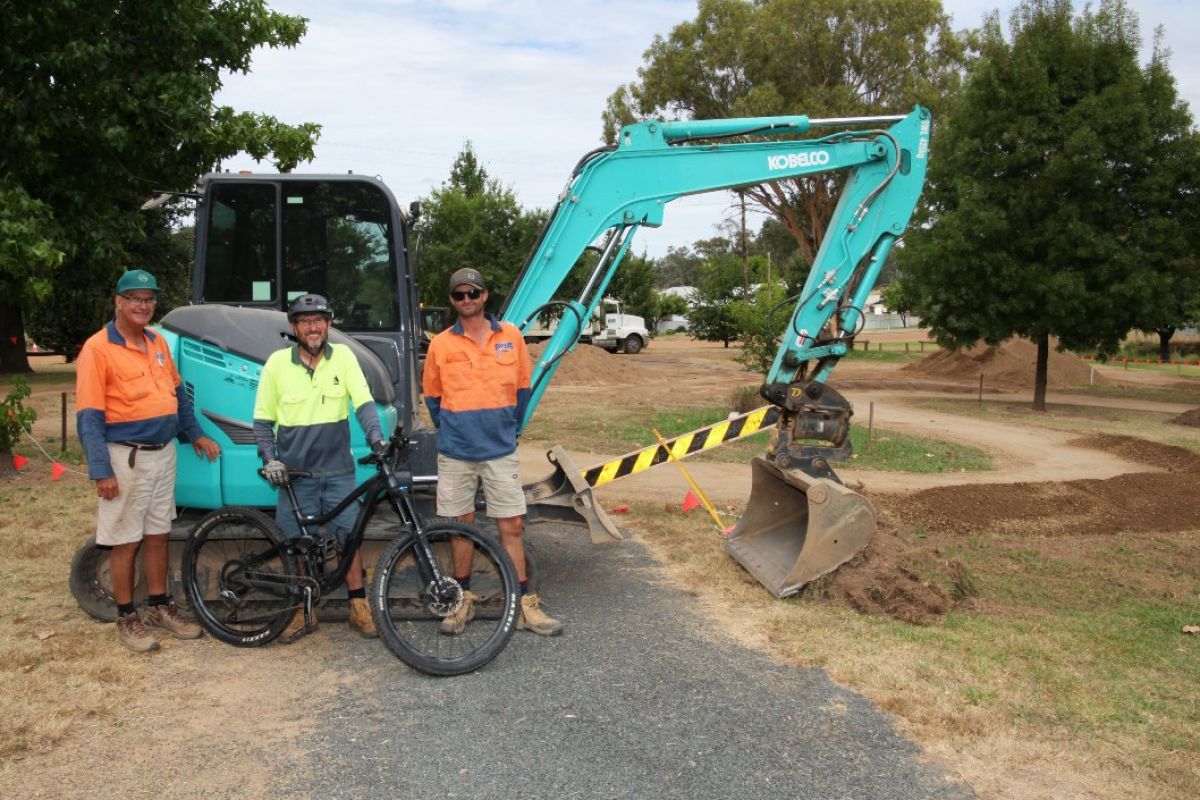 Three men stand in front of an excavator. One of the men has a bike.