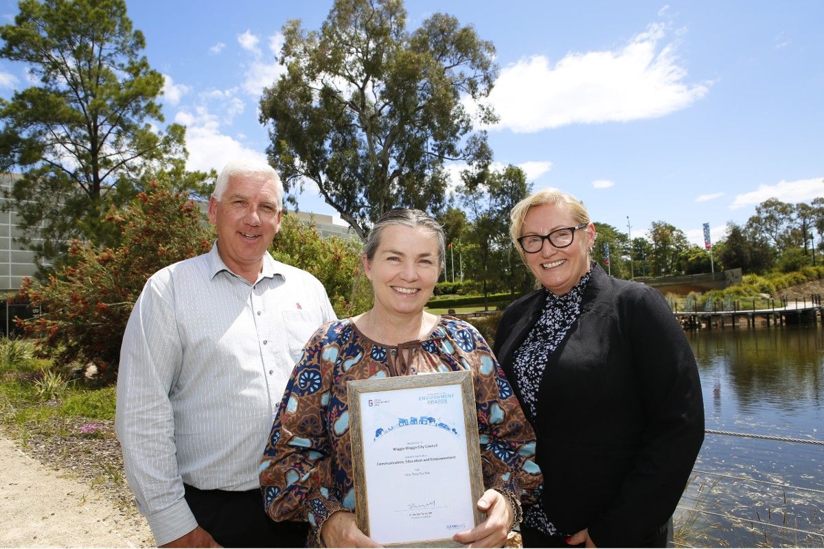 Man and two women, one holding an award, standing beside a lagoon