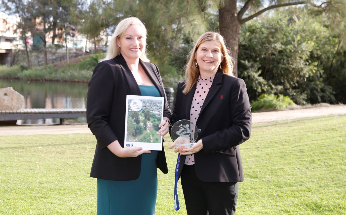 two women standing outside, one holding a document, the other holding a glass trophy. 