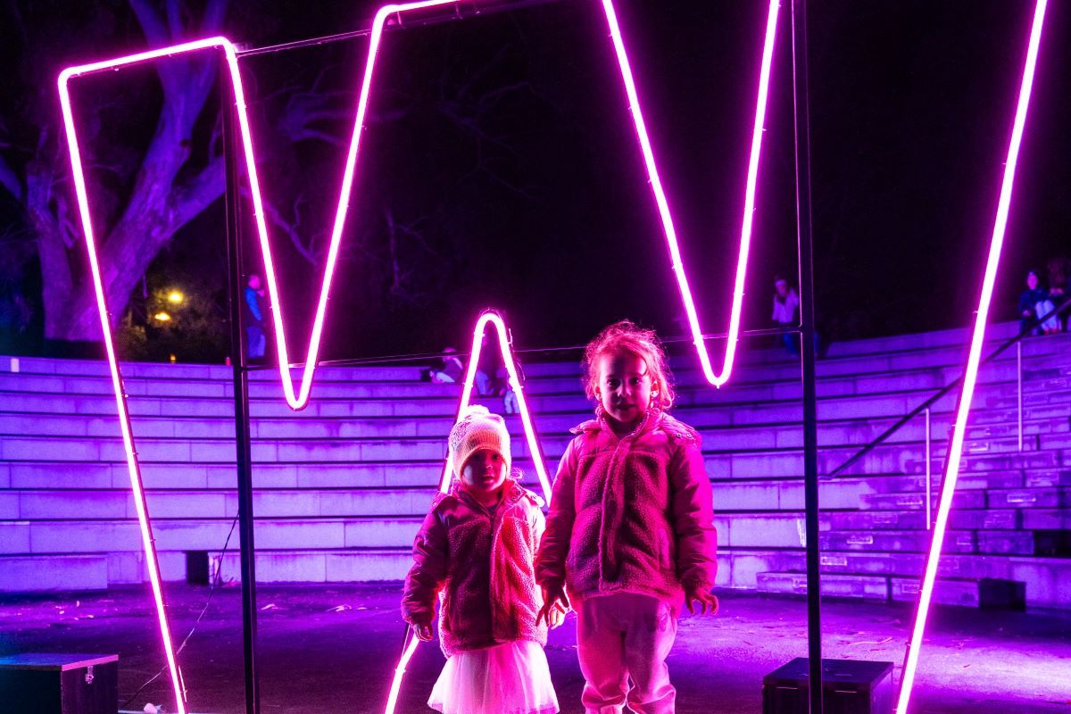 two small children in front of a large pink neon sign in the shape of a W. 