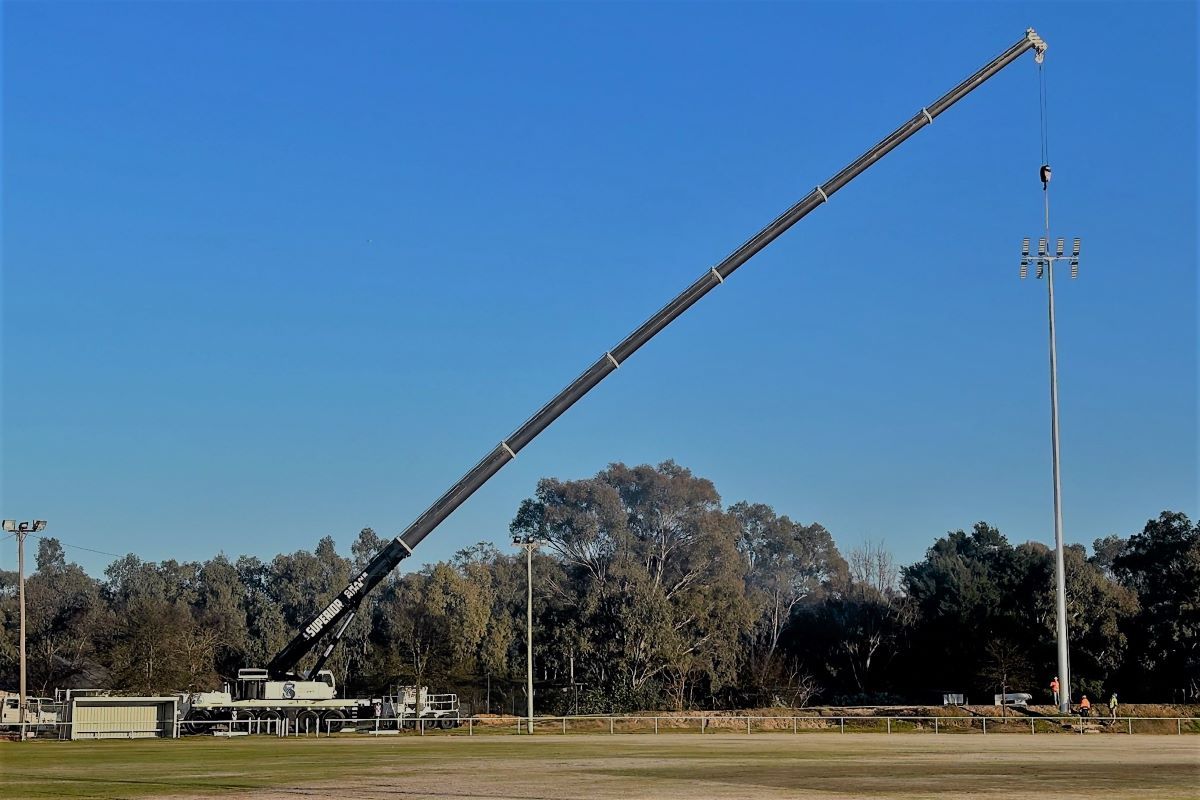 A crane reaches over from left to right, placing a pole in position at a sportsground.
