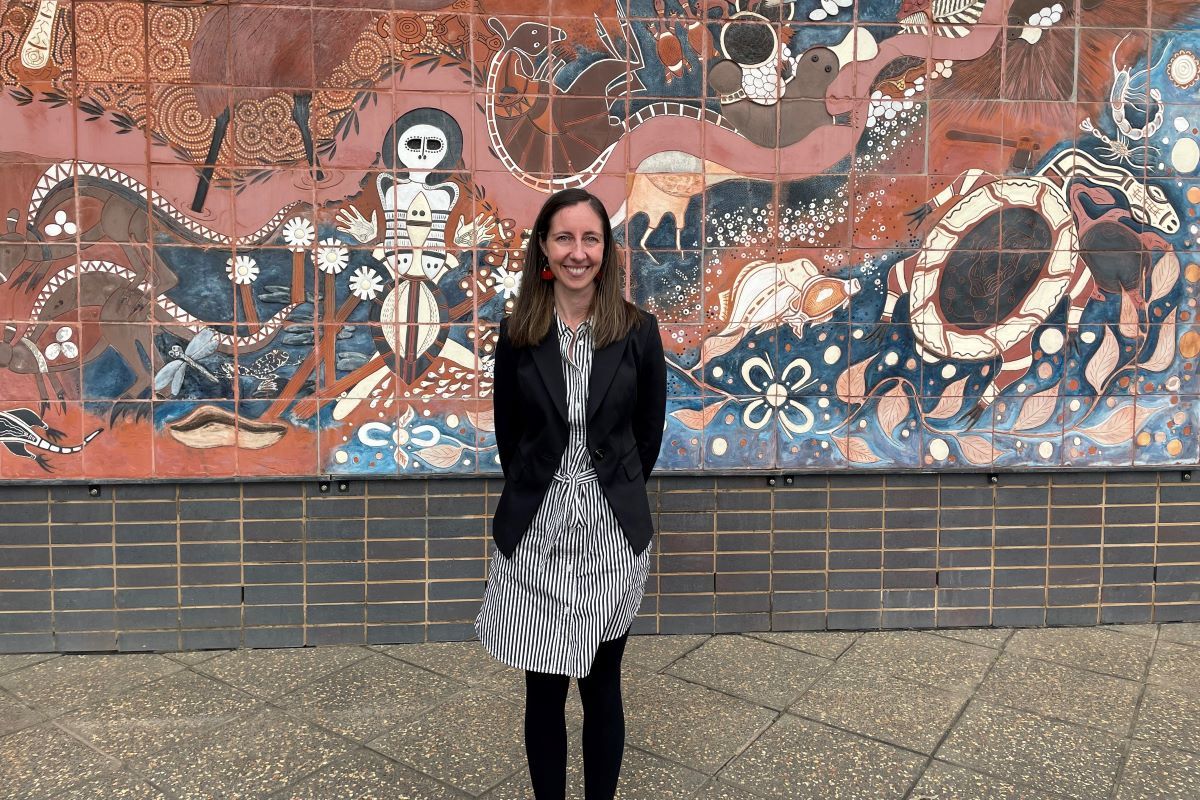 A woman stands for a photo in front of an Aboriginal artwork. 