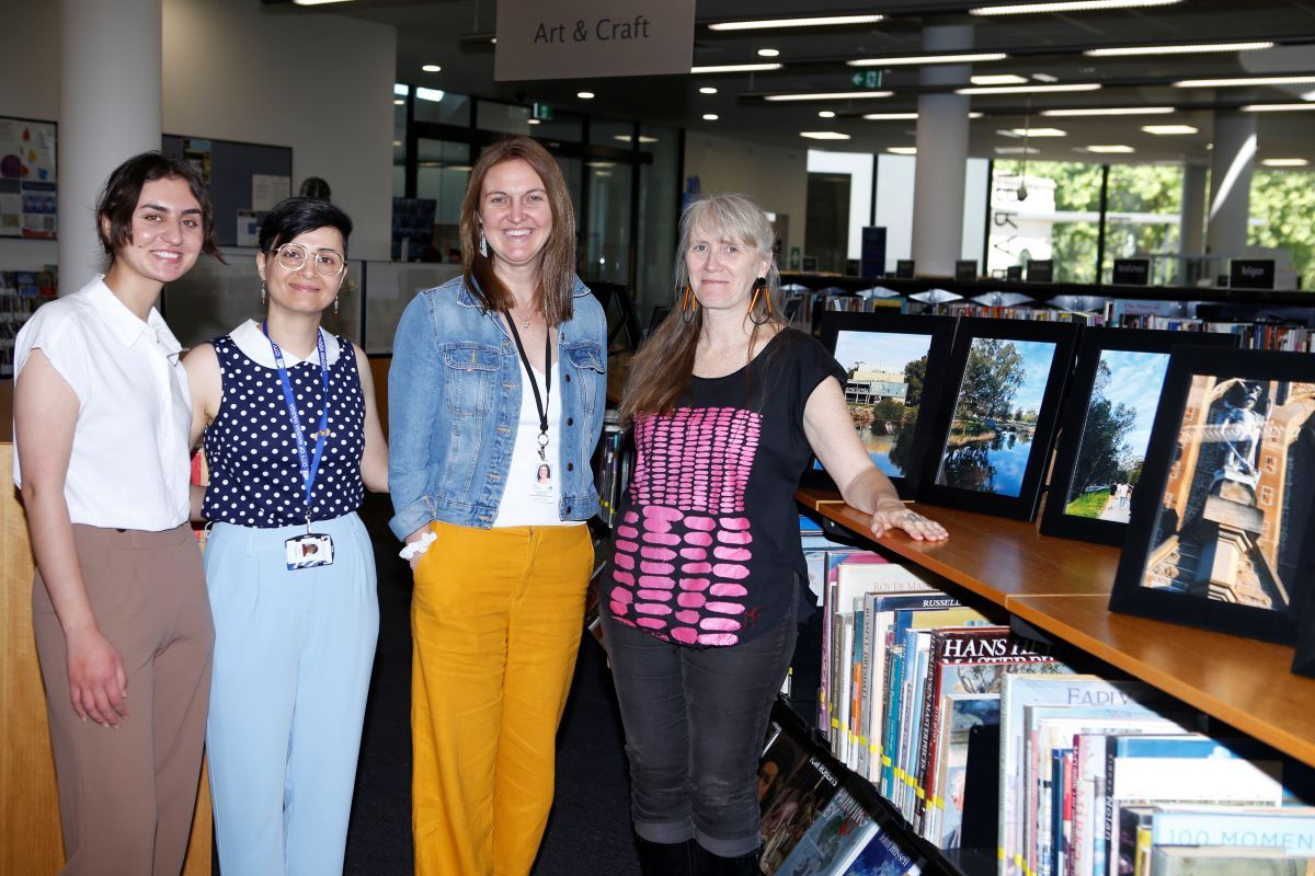 Four women stand together in a public library with a photographic exhibition. 