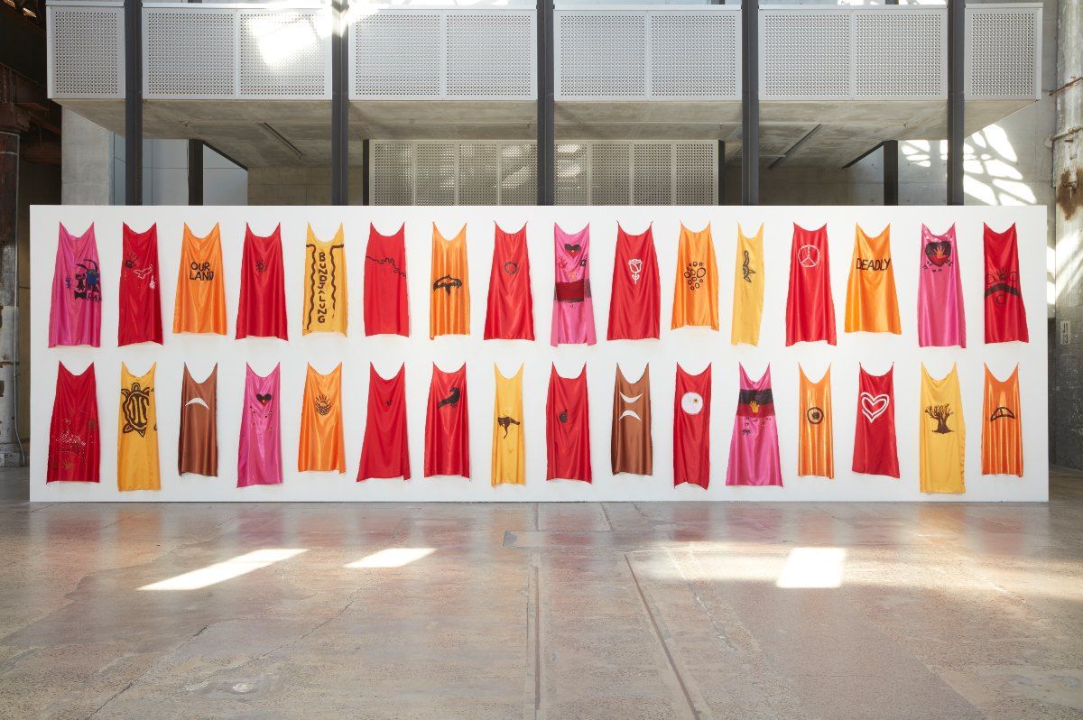 A large rectangular white wall stands tall with sixty capes neatly hung in two horizontal lines along the wall. The capes range from the colours red, yellow, orange and pink. Each cape has a black or white symbol painted on. Each symbol is different and is significant to the artists live experiences and cultural identity.