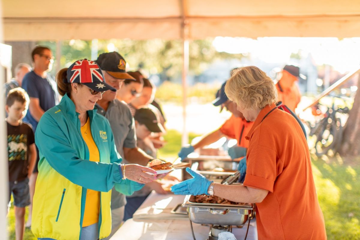 Men, women and children lining up to get served a barbecue breakfast by woman wearing orange shirt