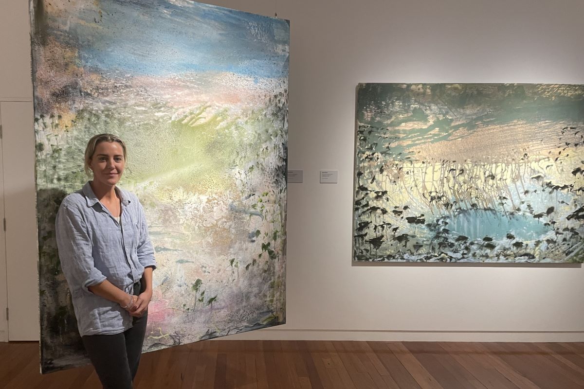 A woman stands in an art gallery space with her large scale landscape paintings. 