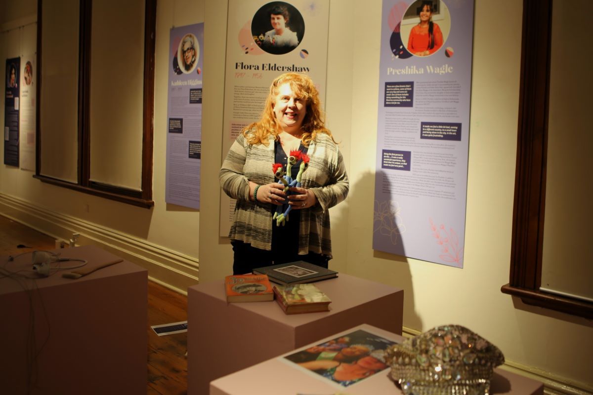A woman stands in a gallery space with items and panels as part of an exhibition about women.