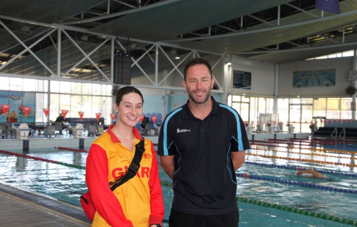 A young woman in a yellow and red lifeguard clothing and a man in black cloth stand beside a public indoor swimming pool. 