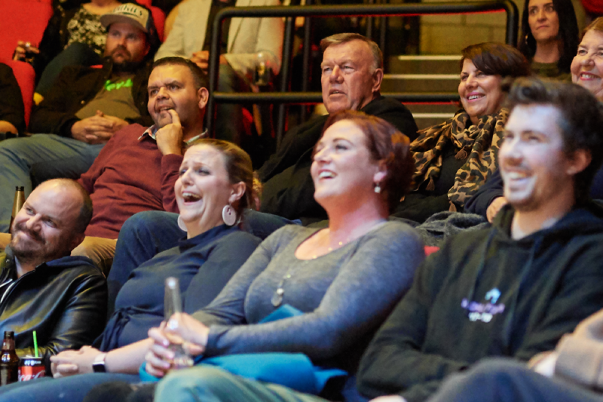 A crowd sitting in a theatre auditorium, laughing and smiling. All are wearing casual attire, some hold drinks in their hands.