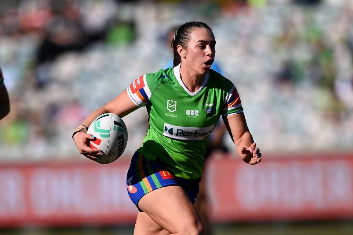 Female Canberra Raiders rugby league player in green jersey running while holding football in right handl.