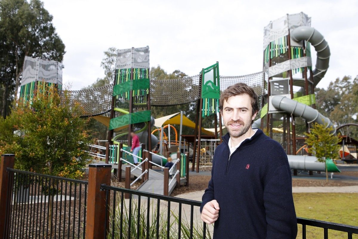 Smiling man with a beard. leaning on a fence with a large playground in the background. 