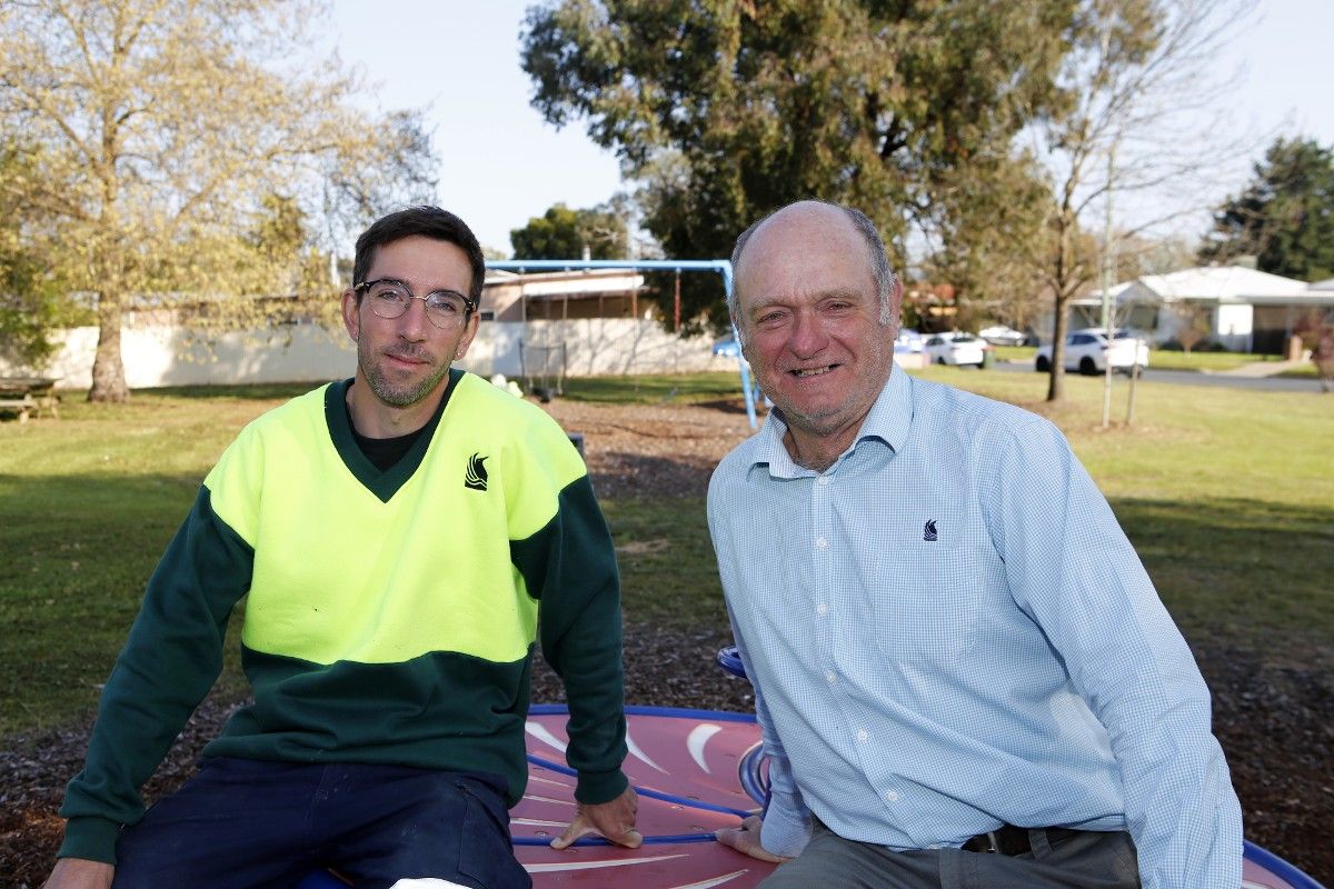 Two men, one in hi-vis jumper, sitting on a spinning wheel at a playground.