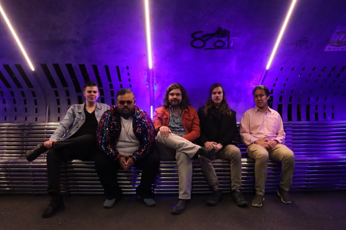 Five funkily dressed men sitting on bench with purple lights above them 