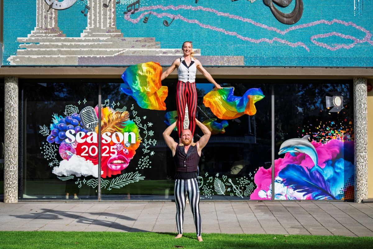 Two circus performers stand in front of the theatre. One performer stands on the shoulders of the other waving rainbow flags.