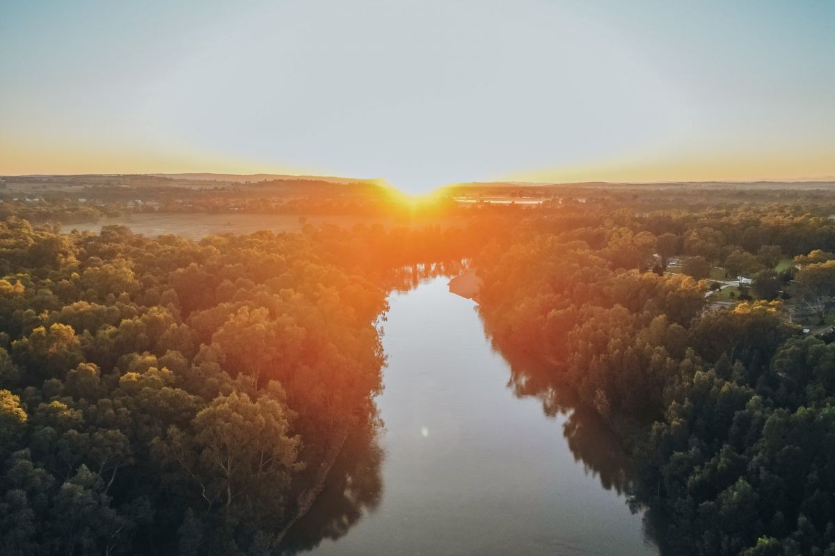 Aerial drone image of river winding through trees, with sun setting on the horizon.