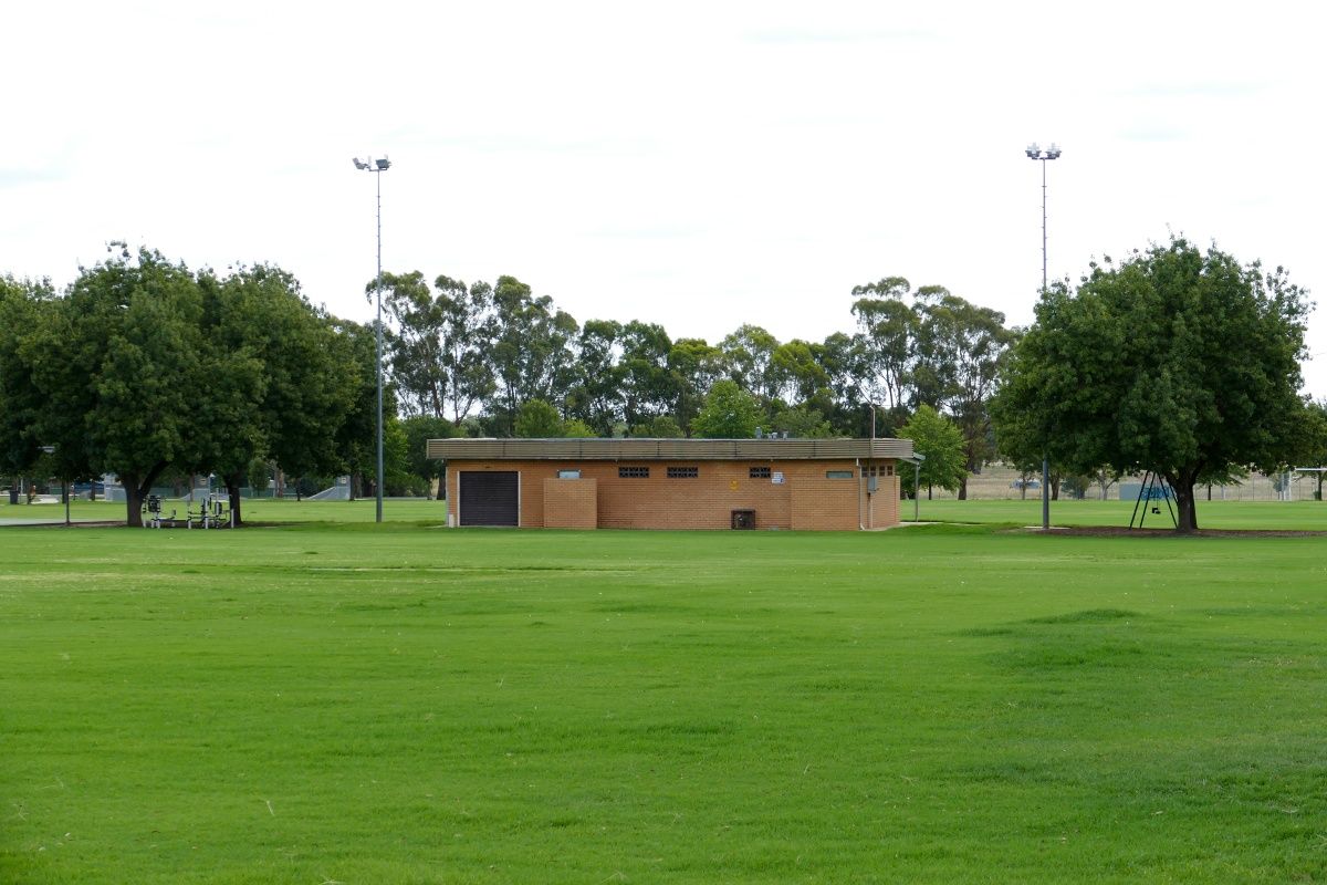 Back view of brick amenities building on a sporting oval, with light towers on either side of the building.