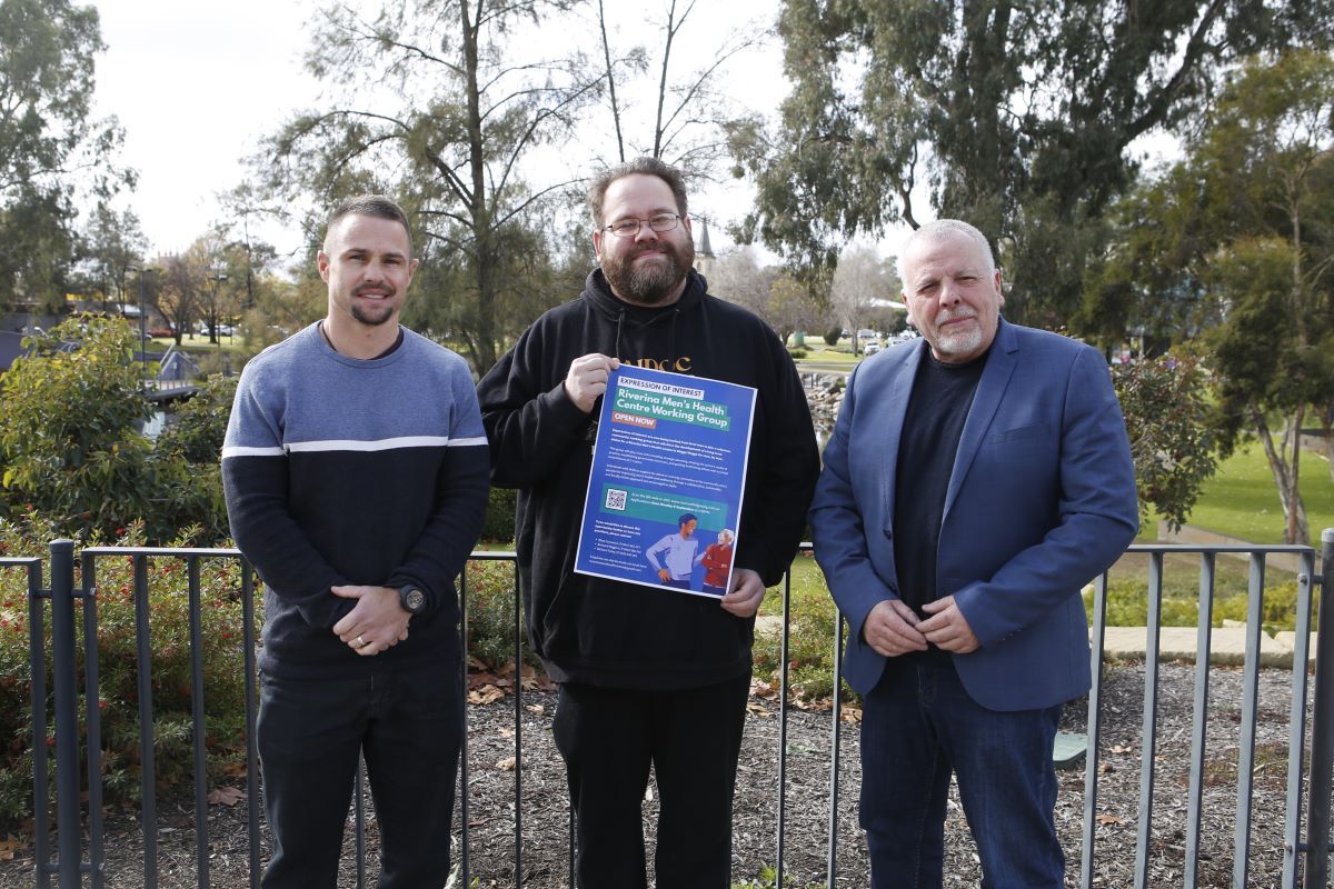Three men standing outside facing the camera, the one in the middle is holding a poster. 