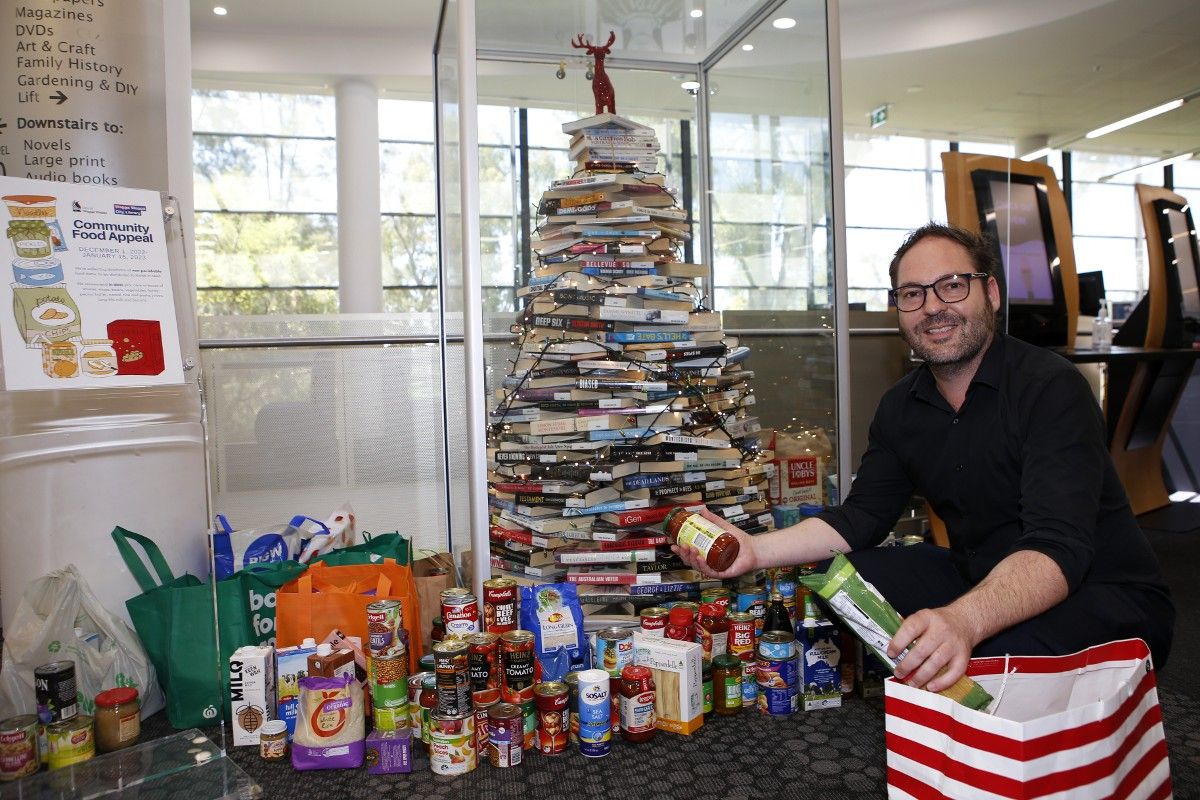 man crouching next to christmas tree, putting non perishable food items at its base