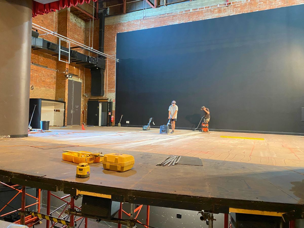 A large stage with no curtains. Two men in the distance are working on it's surface.