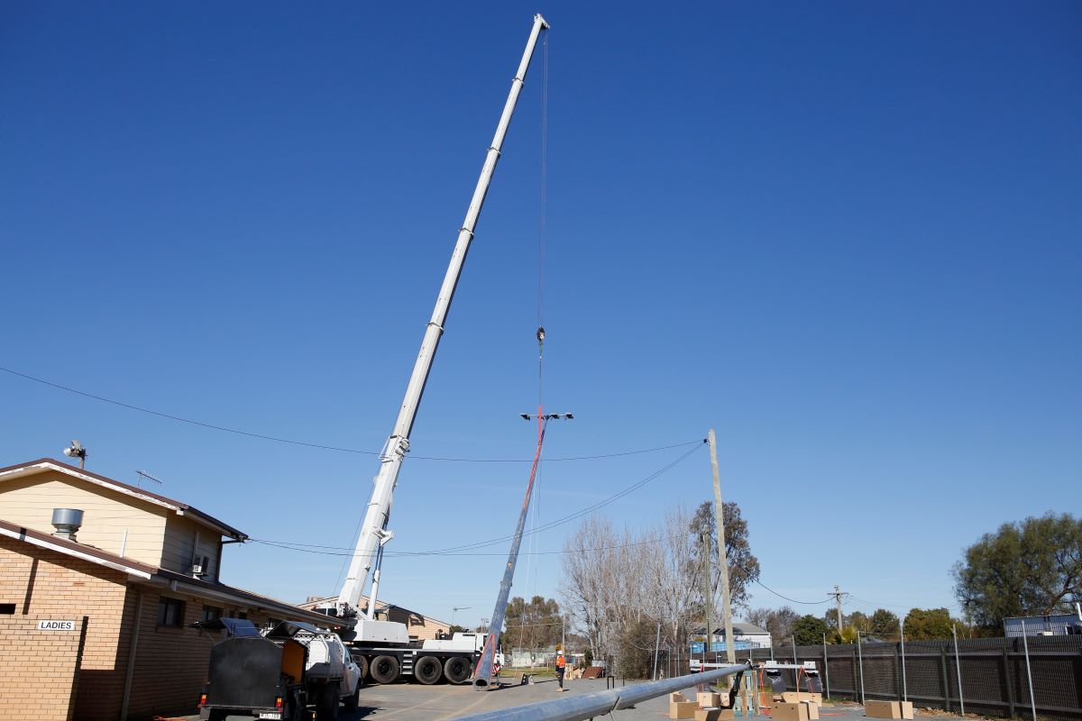 A crane lifts a large light pole to install it at a sportsground.
