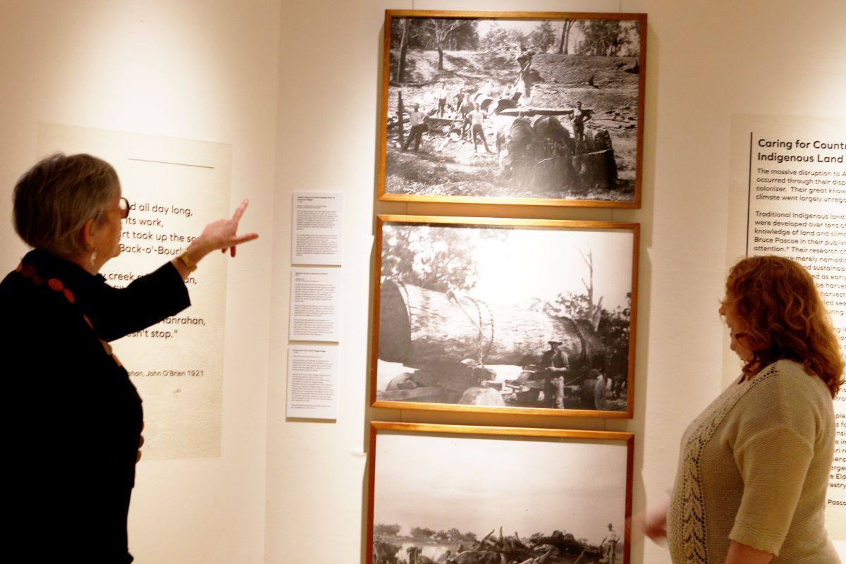 Two women standing in a museum on either side of some black and white historic photographs. 