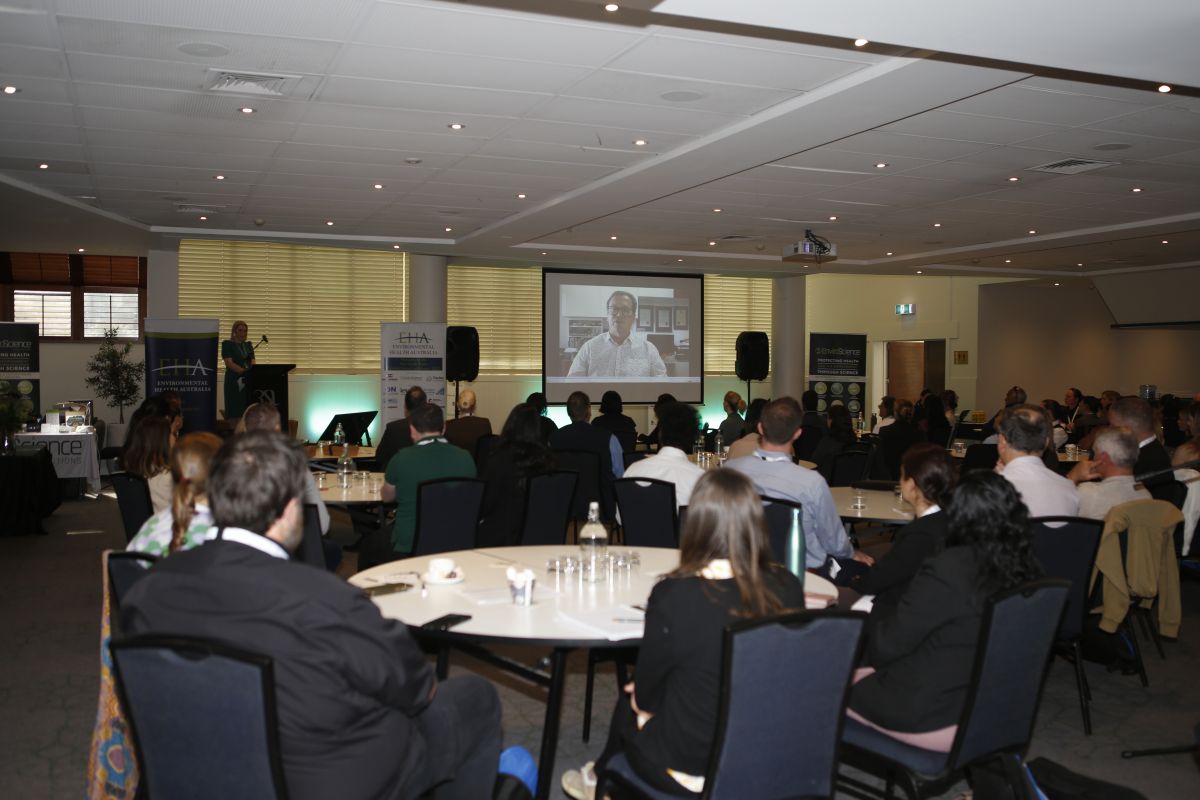 Wide shot of conference. Room downstairs at the RSL with crowd sitting down at tables watching a speech on projector screen.