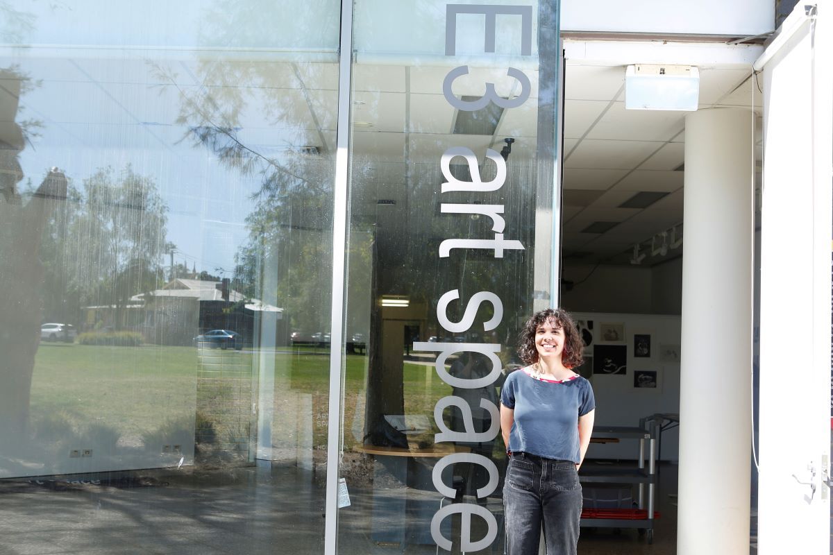 A young woman stands in the doorway of an art studio which has floor to ceiling glass windows, next to the door wording says 