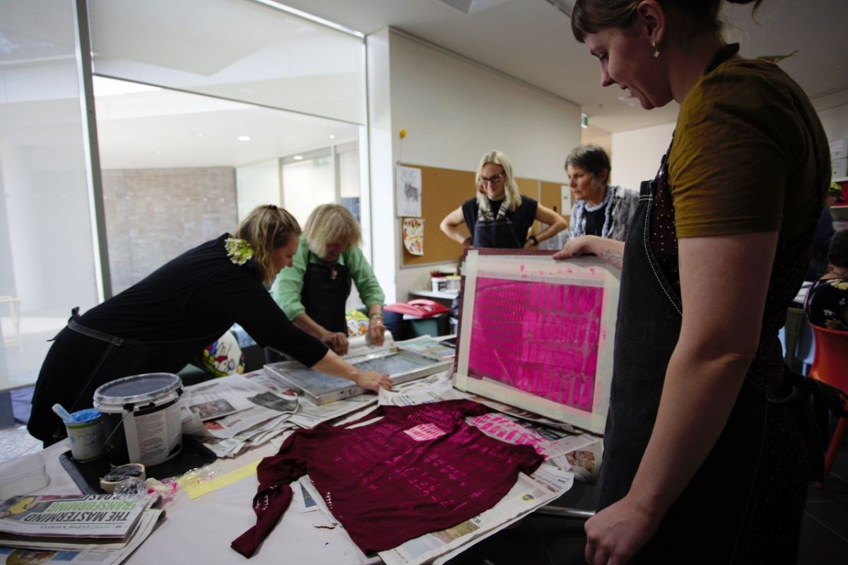 Five women crowd around a table covered in clothing, paint, and other art supplies. The angle is a slight over the shoulder shot of a women printing a paint pattern onto a red top. 