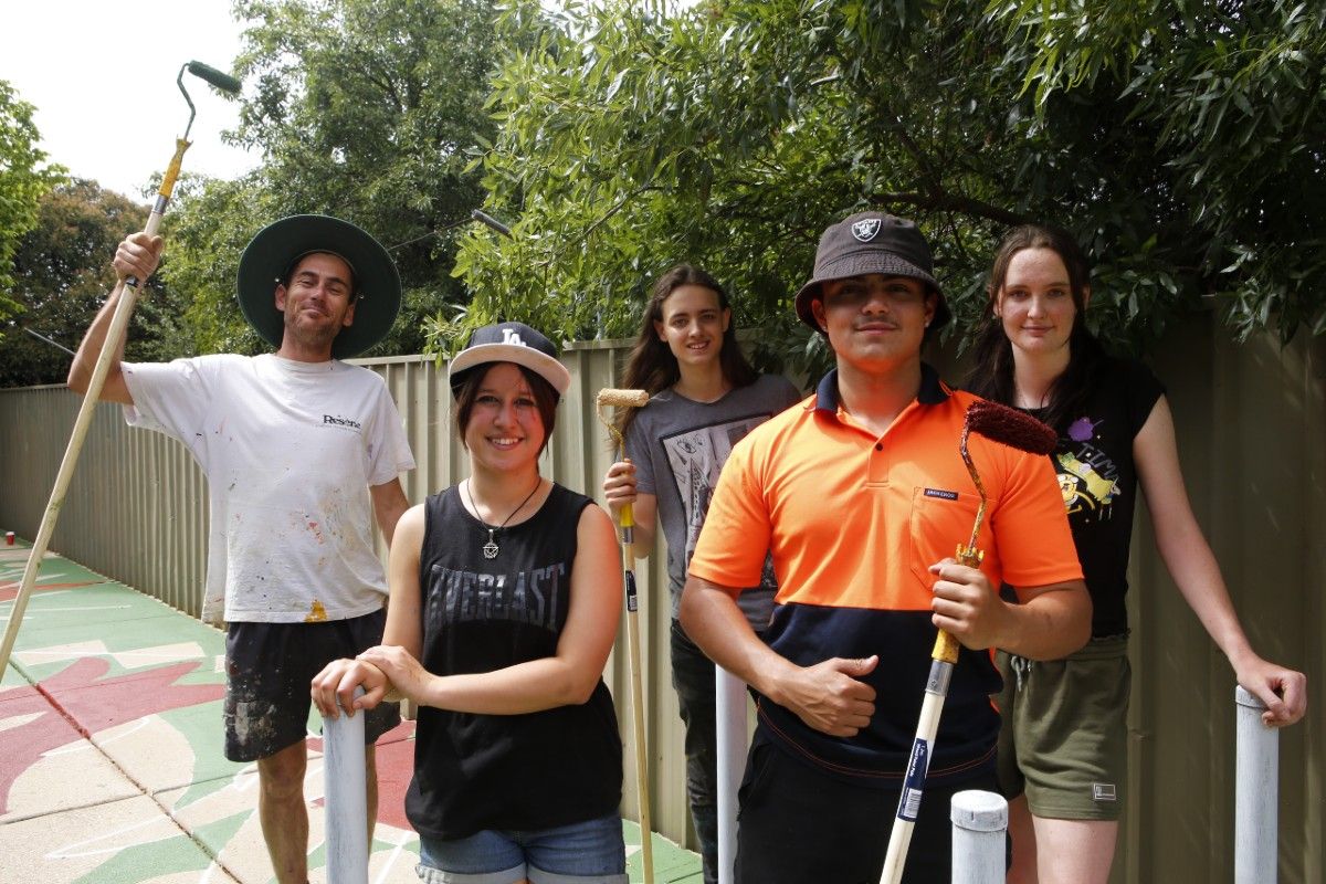 Four students from Mount Austin and artist Damien Mitchell stand in the Mount Austin laneway between Dove Street and Hunter Street. They hold paint rollers and have smiles on their faces. 