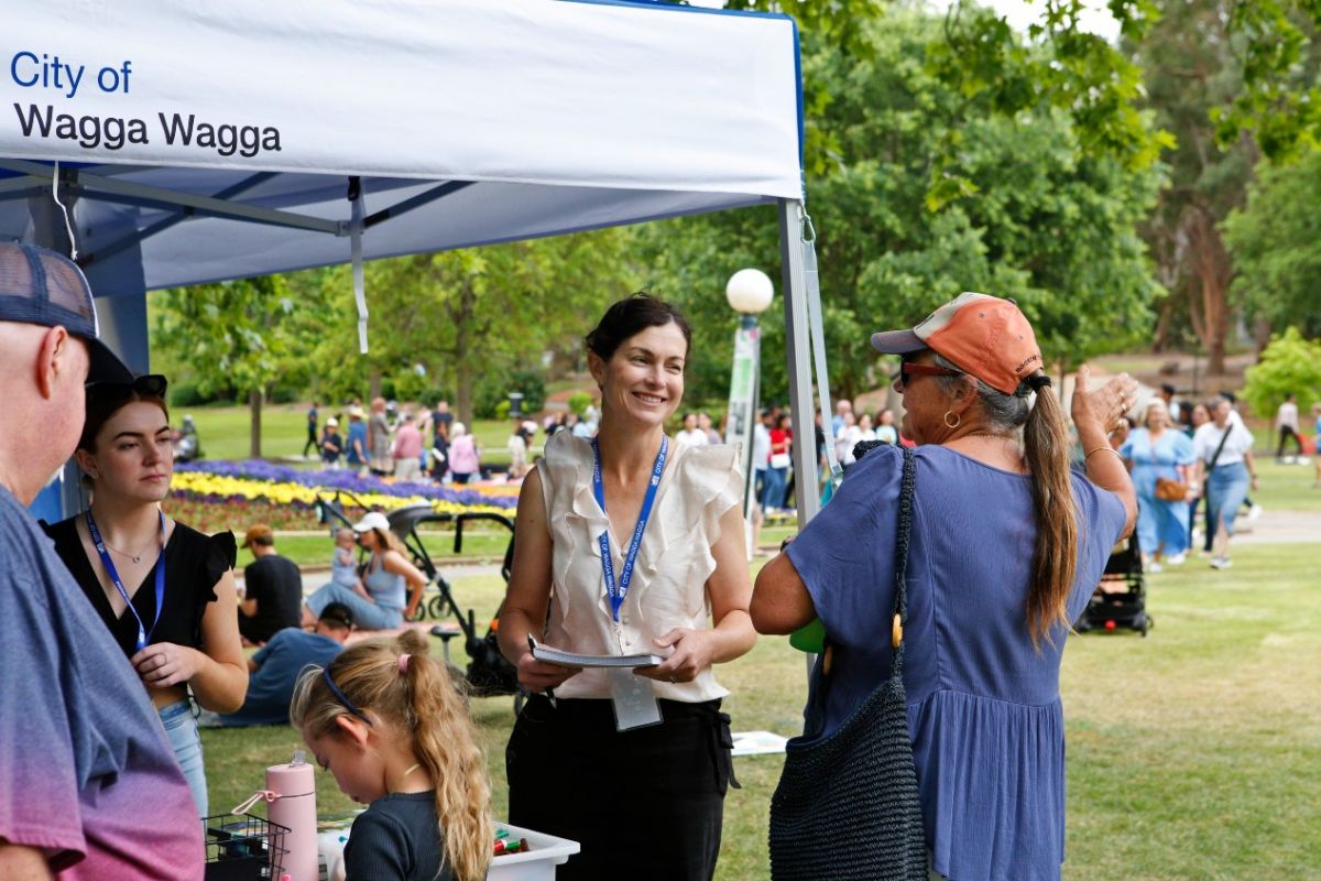 Woman with Wagga Council lanyard talks with family next to City of Wagga Wagga marquee, botanic gardens in background