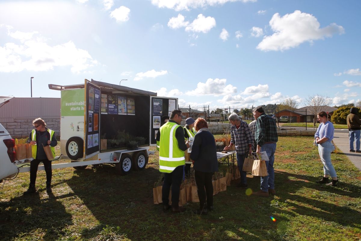 A car, trailer and table is set up on a grassed area.  Seedlings in bags are displayed on the table.. Community members and staff gather around the table.