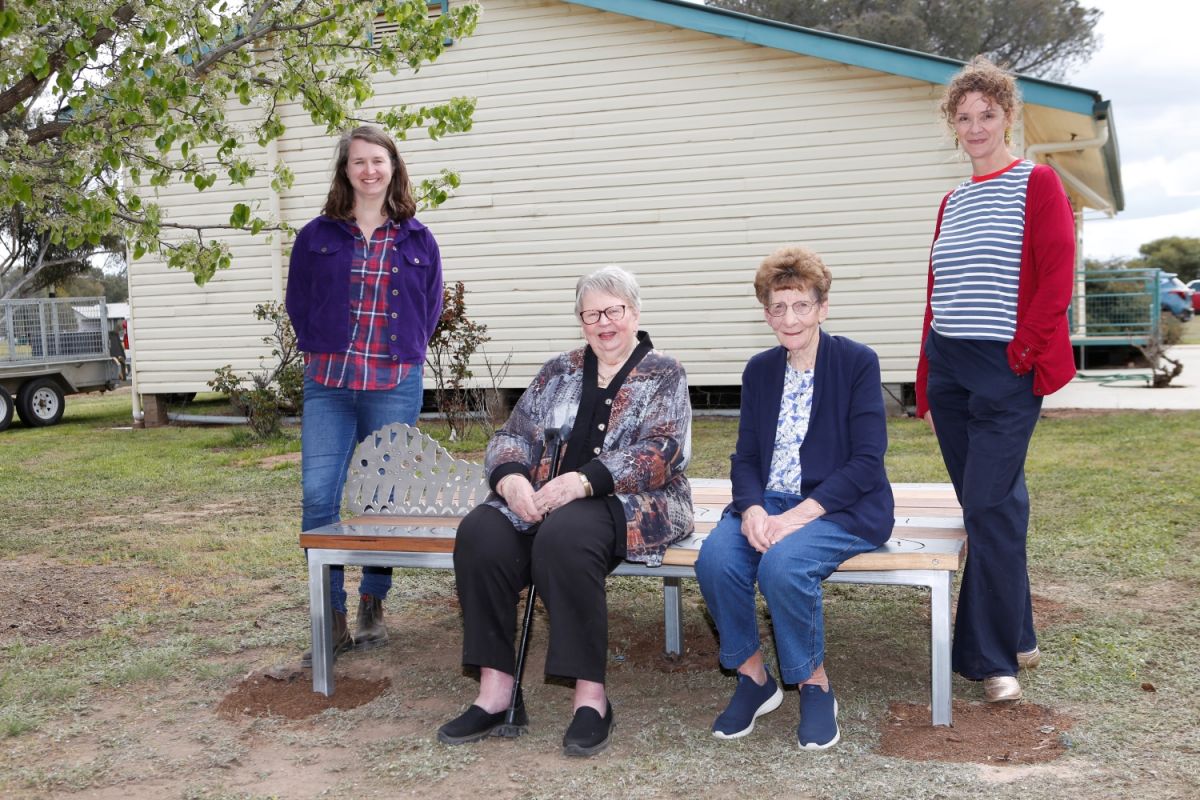 Two women sit on a bench, and two women stand on either side of the bench.