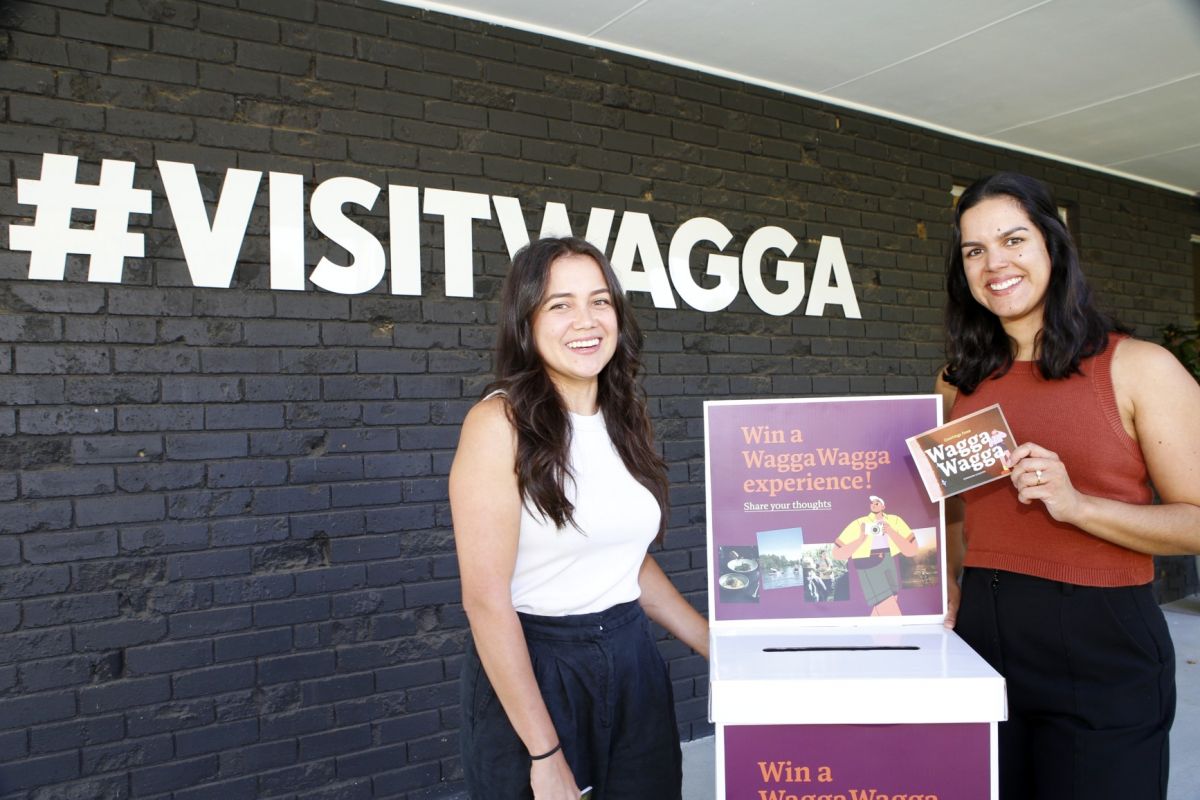 Two women holding promotional material for the Draft Destination Management Plan, standing next to a wall which has a sign reading #visitwagga