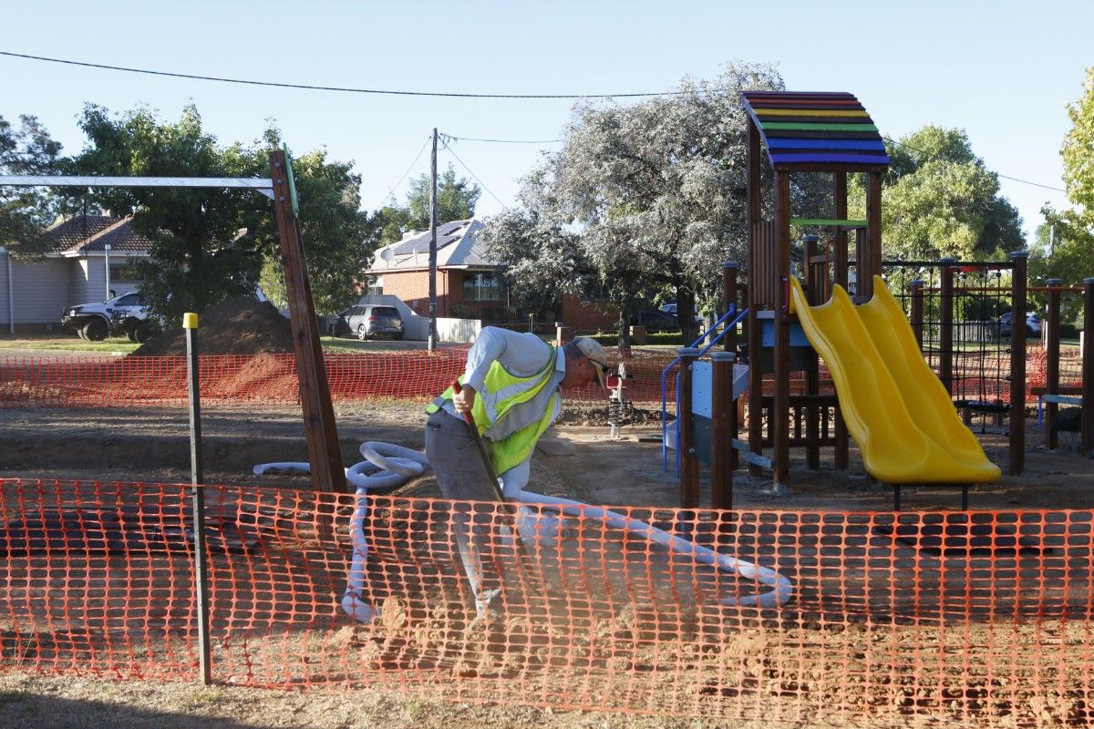 Modular playground tower with slides, climbing net and colourful roof in background, with man in hi-vis vest shoveling soil in the foreground.