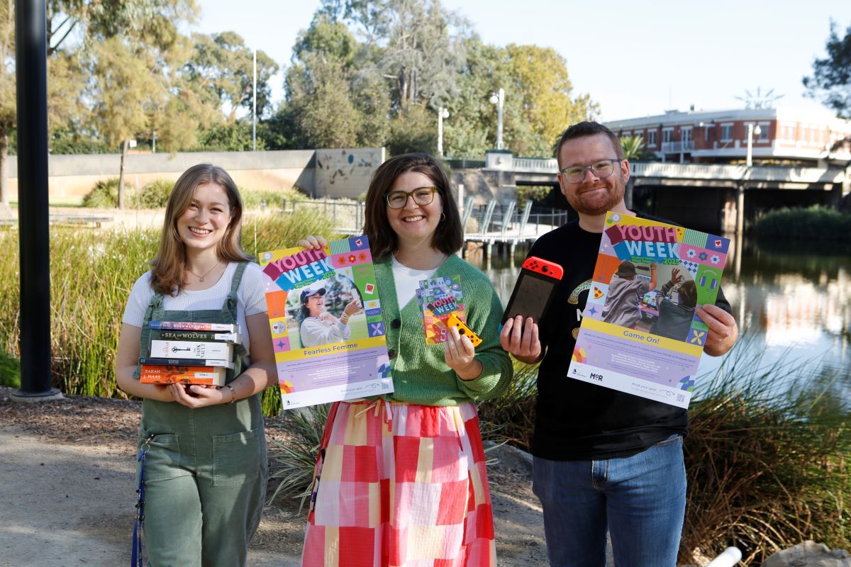 A young woman holding a small stack of books, a young woman holding a poster and info booklet, and a young man holding a Nintendo Switch and poster. They are outside next to the Wollundry Lagoon.