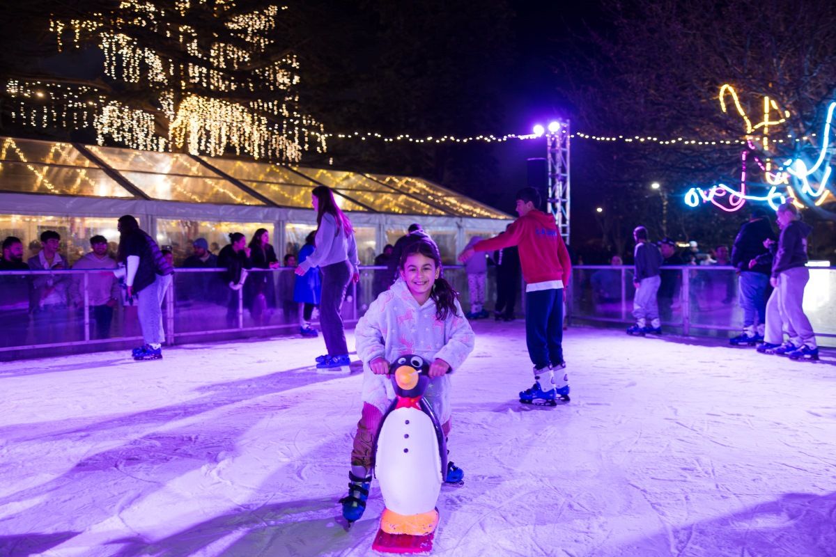 Night time image of a young girl in a colourful winter jacket leaning on a penguin skating as she skates around an ice rink, with other people skating in the background.