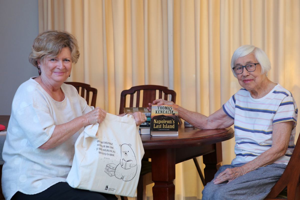 Two women sitting at table with stack of books and bag, containing books