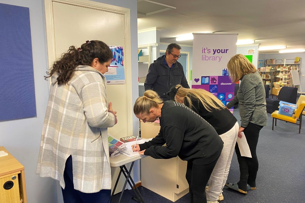 Three woman leaning over signing paperwork, assisted by agile library officers