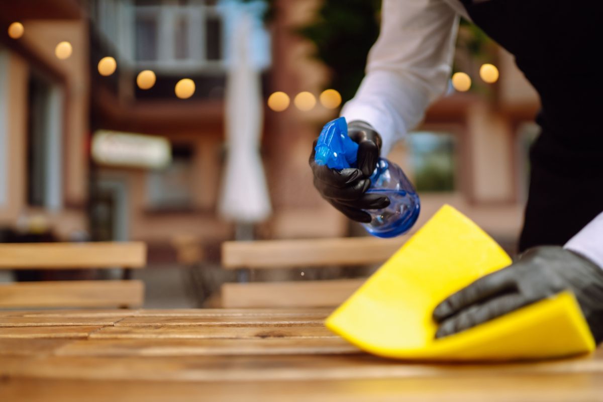 Wooden table being cleaned by person wearing black gloves, holding a spray bottle and wash cloth