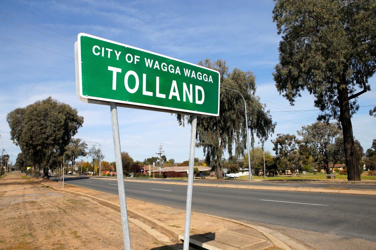 A green suburb sign with white writing on the side of a road in a suburban area. The sign says 