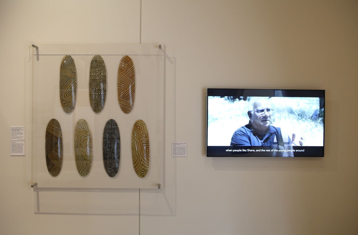 Some First Nations contemporary shields on a wall in a museum, next to screen with a man speaking. 