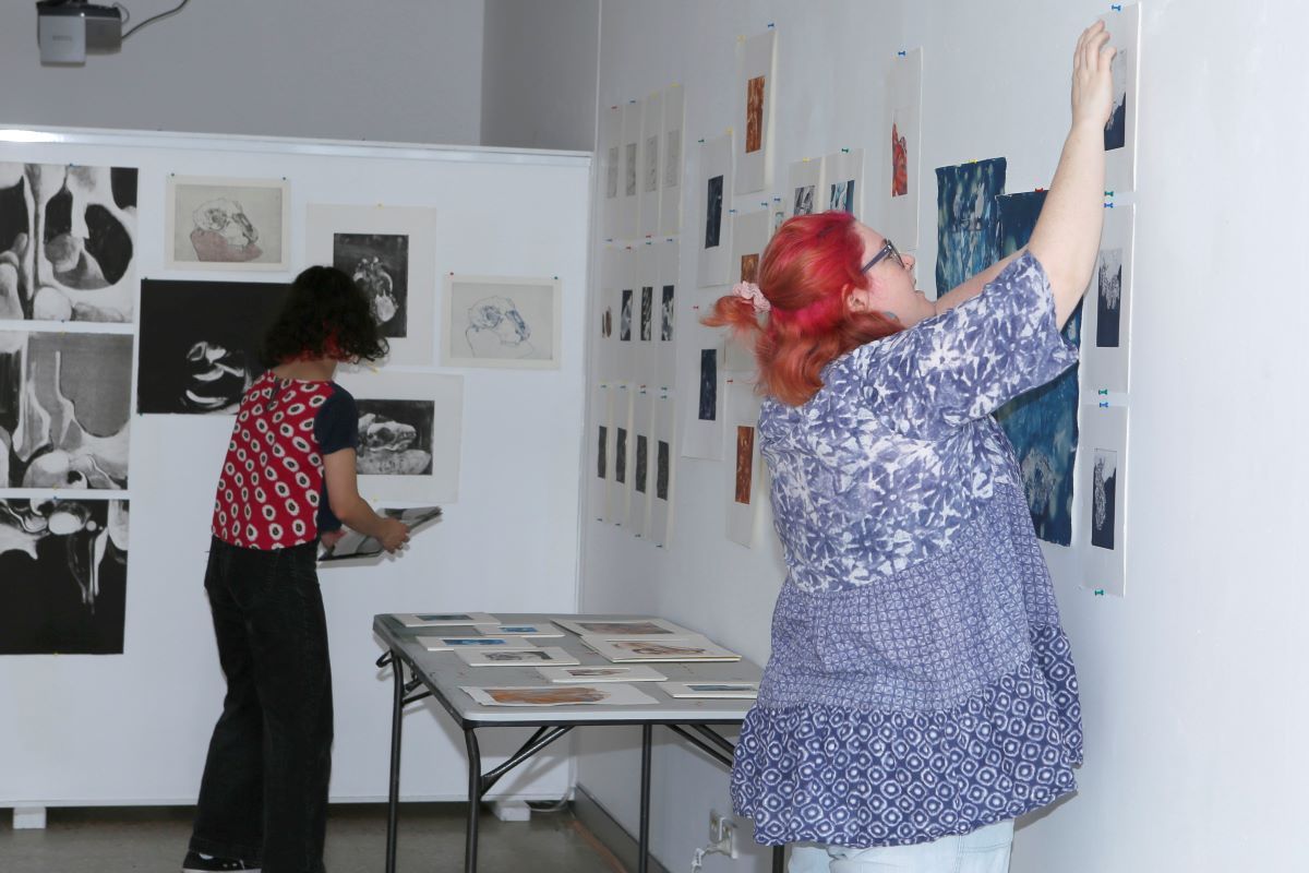 A young woman pins some artworks on paper on the wall of a gallery space. Another woman in the background is sorting out artworks.