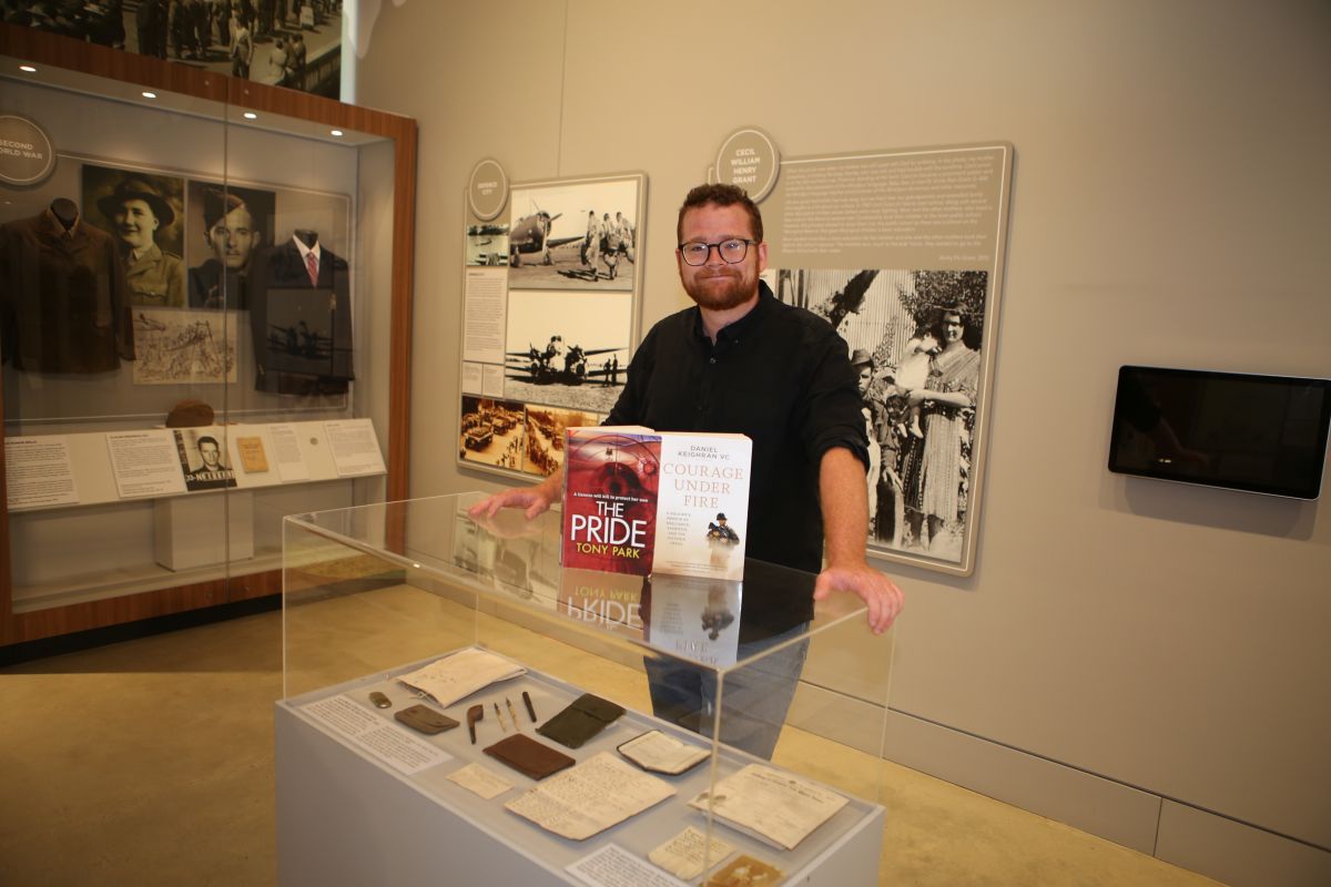 A man standing in a museum setting with two books sitting on a display cabinet. 