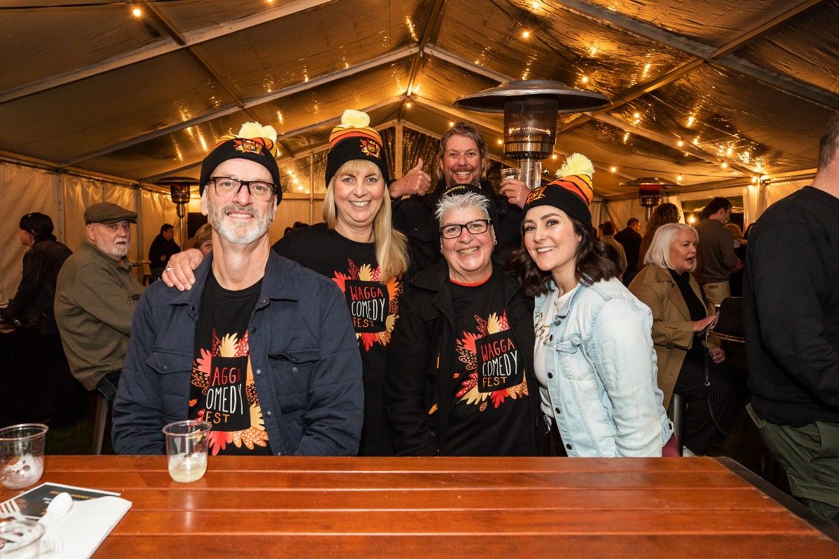 Group of men and women wearing Wagga Comedy Fest t-shirts, sitting at table under a marquee, with other people in the background.