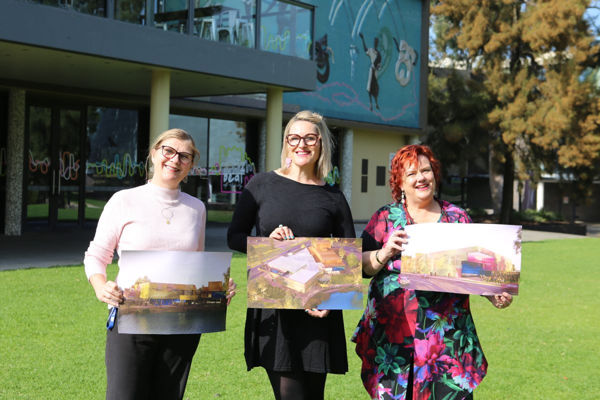 Three women standing in front of a theatre building holding concept designs for a new performing arts centre.