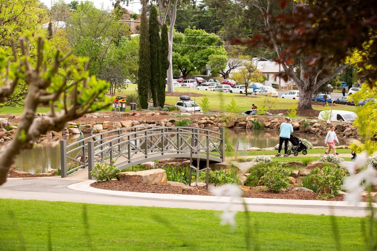 Family walking through gardens next to pone, with pedestrian bridge in foreground.