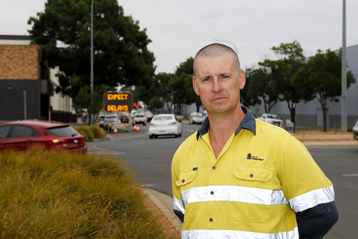 Man in a hi-vis shirt stands on the footpath with a roundabout and road in a CBD area in the background, along with an electronic message board stating ‘expect delays’.